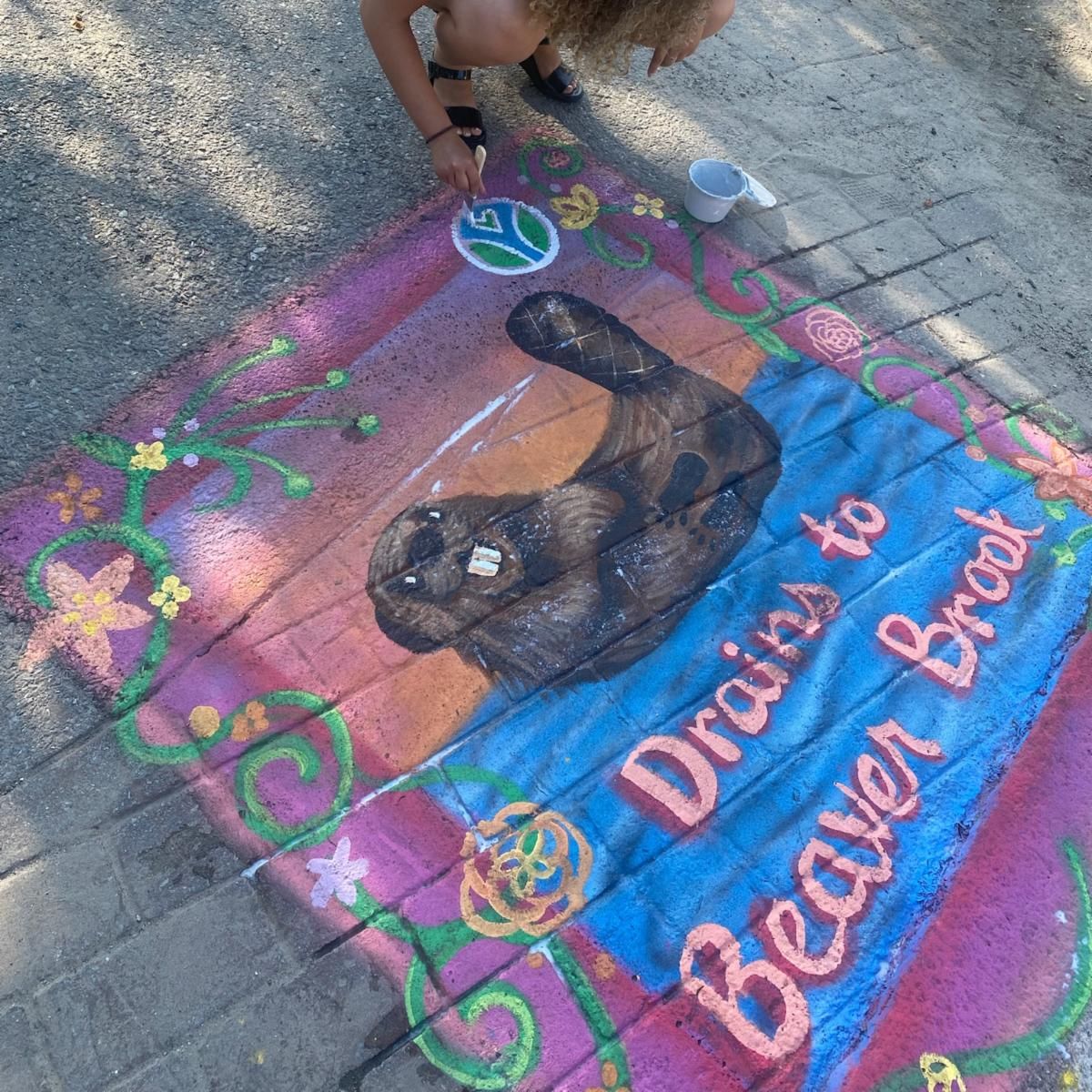 Jennessa Burks painting a colorful mural on a sidewalk featuring a realistic beaver swimming, surrounded by vines and flowers. The mural reads “Drains to Beaver Brook.”