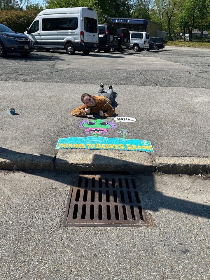 Maggie Grenier lying next to a finished mural featuring a green cartoon monster rising from water holding a six-pack ring. A speech bubble says “Bruh…” with the message “Drains to Beaver Brook.”