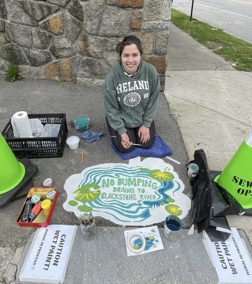 Ellie Ellis kneeling next to a freshly painted mural of a blue-green water scene with lily pads and frogs. Text reads “No Dumping – Drains to Blackstone River.” Supplies and cones surround the site.