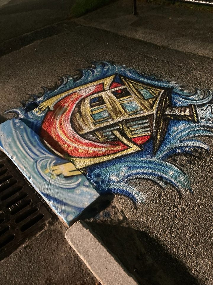 Storm drain painting of a yellow triple-decker cradled in a red life preserver, surrounded by dynamic blue waves at night.