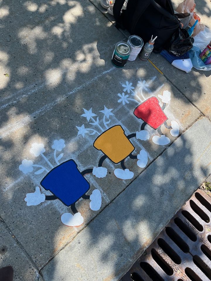 Partially painted mural of three colorful cartoon flower pots (red, yellow, blue) with smiling faces and walking legs. The flowers are outlined in white. The mural is being painted next to a storm drain, with painting supplies and a backpack nearby.