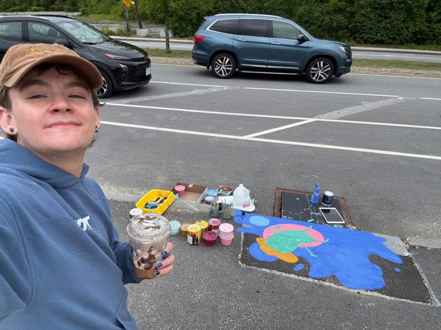 Caoin O'Durgy smiling with a drink in hand in front of a partially painted storm drain mural of a pink and green turtle on a blue background. Art supplies, paints, and brushes are scattered on the pavement.