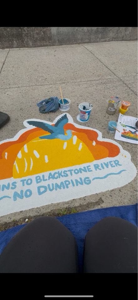 Storm drain mural featuring a blue heron flying over a marsh at sunset. The nearly completed text reads “Drains to Blackstone River – No Dumping.” Paint cups and a reference sketch are nearby.