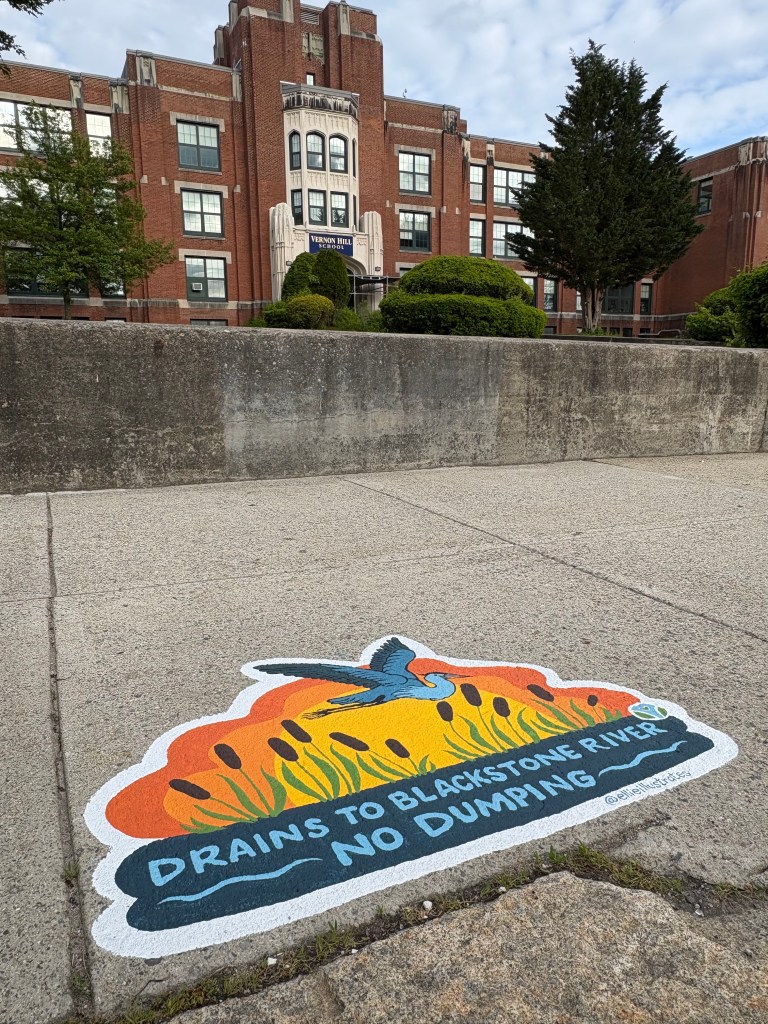 Storm drain mural in front of Vernon Hill School showing a blue heron flying over cattails with a vivid orange and yellow sunset in the background. The mural reads “Drains to Blackstone River – No Dumping.”