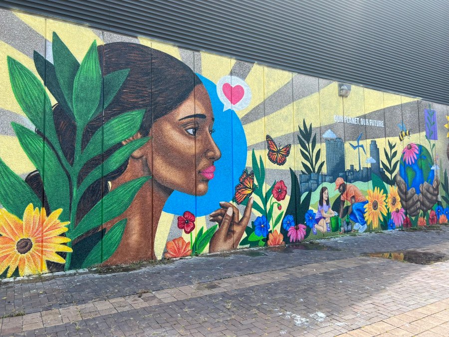Full mural featuring a woman’s face surrounded by flowers, butterflies, and community scenes with people gardening and caring for nature. The background shows the city skyline and a banner that reads “Our Planet, Our Future.”