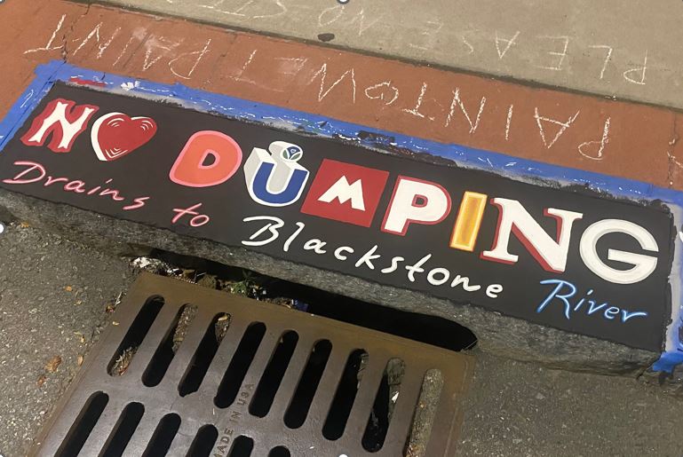 Sidewalk mural with a black background and multicolored, stylized letters spelling “NO DUMPING.” Beneath it, cursive red and white text reads “Drains to Blackstone River.”