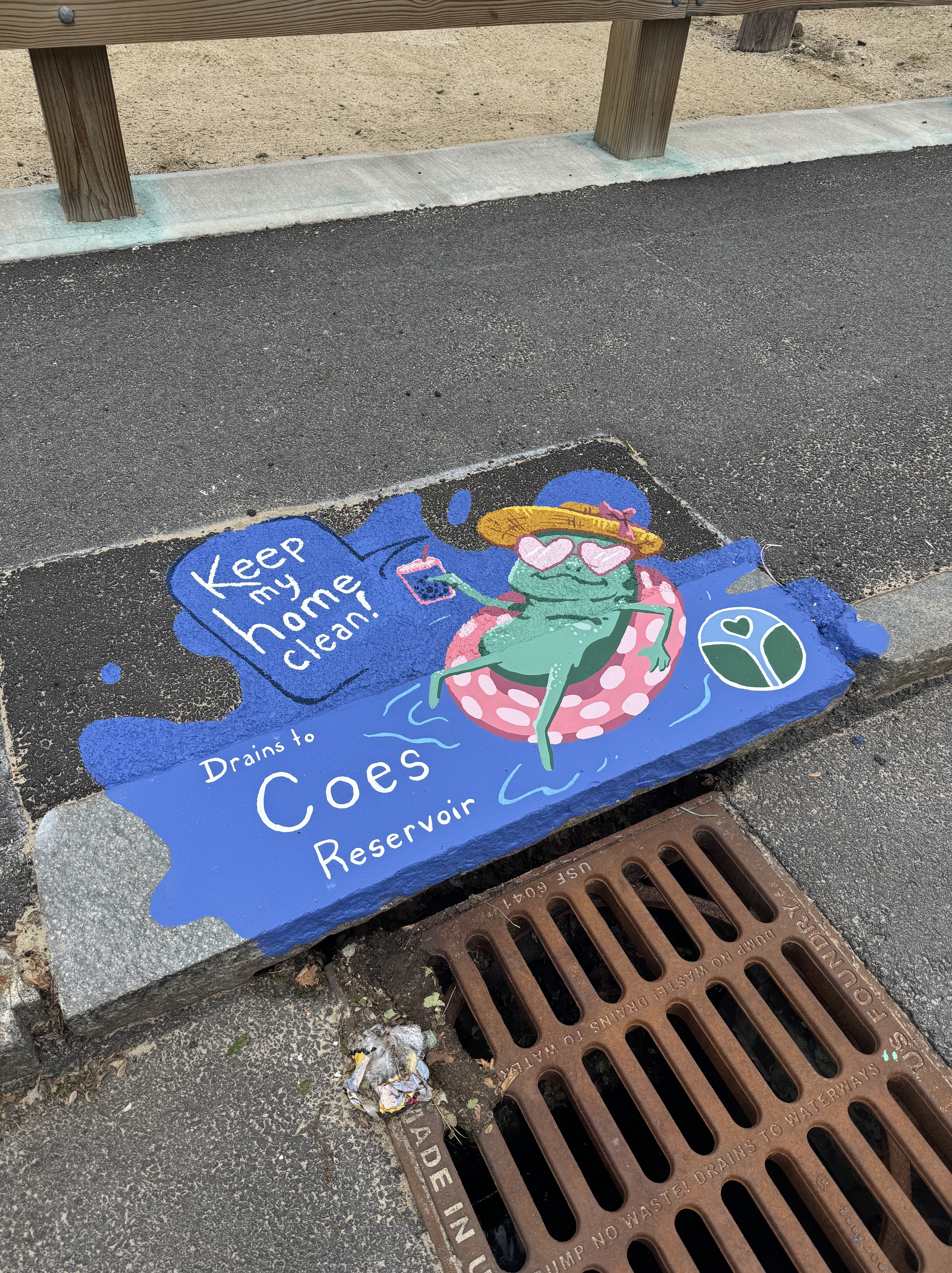 Sidewalk painting of a relaxed frog wearing heart sunglasses, sipping a drink while floating on a pink polka-dot inner tube. The message reads: “Keep my home clean! Drains to Coes Reservoir.”