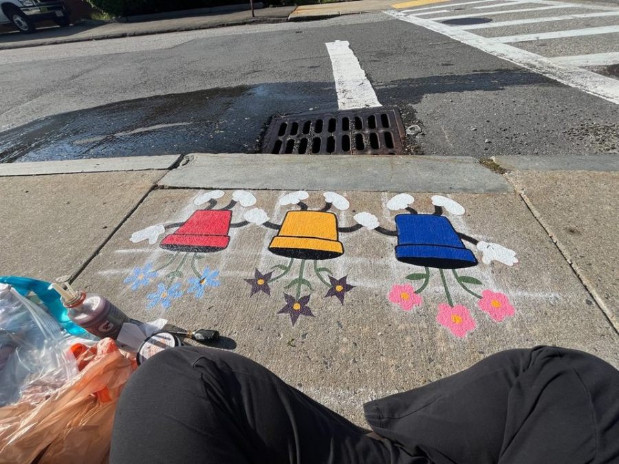 Initial stage of the mural showing three flower pots in red, yellow, and blue, each with flowers. The faces and limbs are not yet filled in. The mural is painted along a sidewalk near a street corner and storm drain. Paint cans and supplies are visible in the corner.