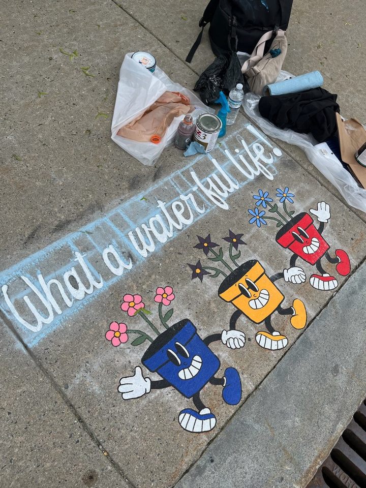 Partially completed sidewalk mural featuring three colorful cartoon flower pots with arms and legs. The pots are blue, yellow, and red, with smiling faces and blooming flowers. The phrase “What a water-ful life!” is sketched in white chalk above the design. Paint supplies are scattered nearby.