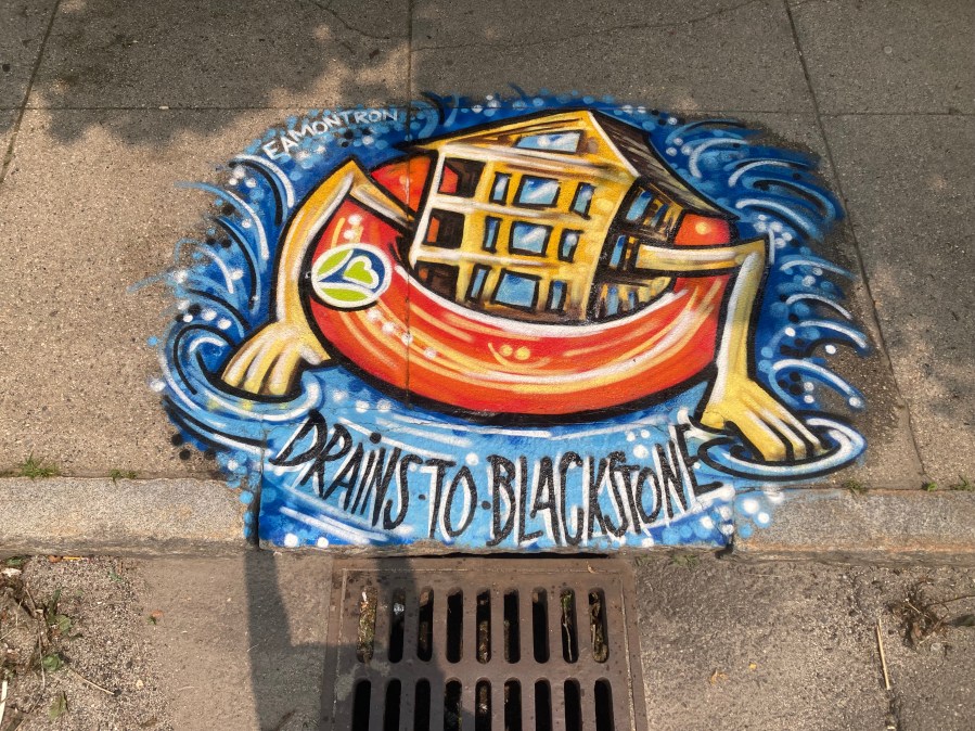 Full view of a mural showing a yellow triple-decker house with three stacked porches and three windows per floor, floating in a red and yellow inner tube. The house has playful cartoon arms, and the water beneath it flows toward a nearby storm drain. The mural includes the message “NO DUMPING – DRAINS TO BEAVER BROOK.”