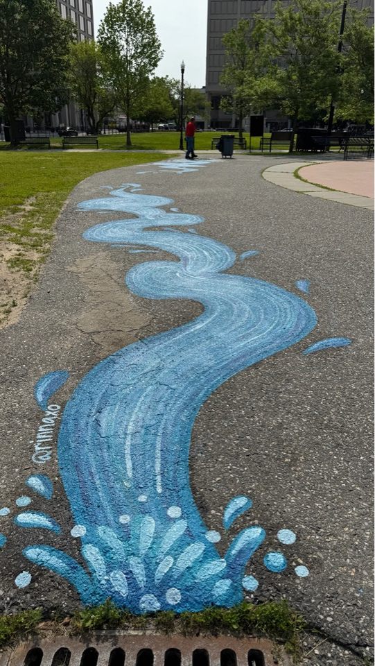 Large painted mural of a winding blue stream flowing toward a storm drain, creating the illusion of water being funneled into the drain. The stream curves down a park walkway surrounded by grass and trees.