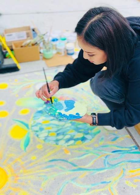 Artist Aiko Itadani is crouched over a sidewalk mural in progress, carefully painting with a brush in one hand and a palette of blue and green tones in the other. The mural, titled "(Circulation)," features a stylized green turtle with a leafy shell, floating through flowing water among bright yellow suns and swirling plant life. Around her are jars of paint, brushes, and tools. The scene captures a quiet, focused moment in the mural’s creation, highlighting Aiko’s attention to detail and the vibrant, interconnected theme of water and life.