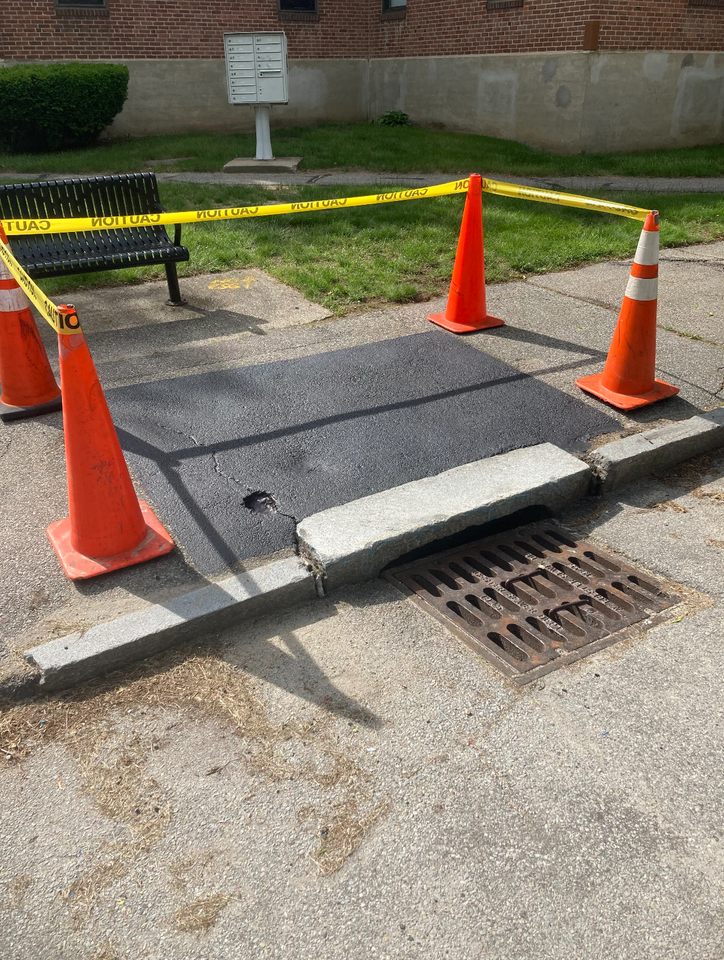 Photo of a paved sidewalk next to a storm drain where the “3 Decker Lounging” mural once stood. The mural, which featured a yellow Worcester triple-decker floating in an inner tube, was sadly painted over before the artist could complete the final details.