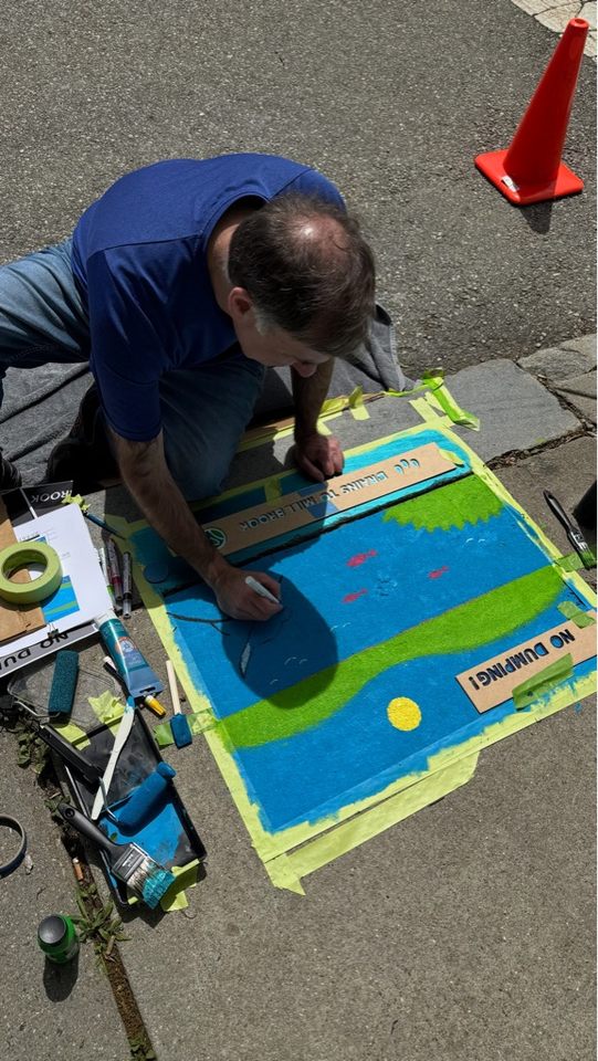 Artist Greg Moutafis sitting on the sidewalk while painting a colorful storm drain mural. The design features a blue river scene with green hills, a yellow sun, red fish, and a black and white bird on a branch. Stencils and painting tools are scattered nearby.