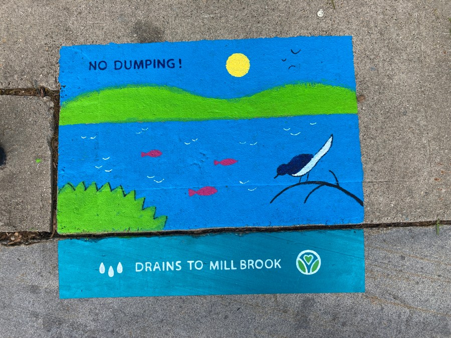 inished storm drain mural showing a bright blue river with red fish, green hills, a yellow sun, and a bird perched on a branch. The text reads “NO DUMPING! DRAINS TO MILL BROOK.” Three water droplet icons and Worcester Green Corps's heart-shaped Earth symbol logo are also included.