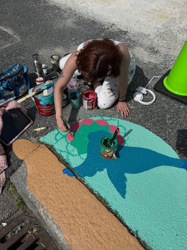 Artist Colomba Klenner kneeling on the pavement while painting the background of a storm drain mural. The design features the outline of a blue seal with a pink and green mandala behind it. Paint cans and brushes are scattered around the artist.