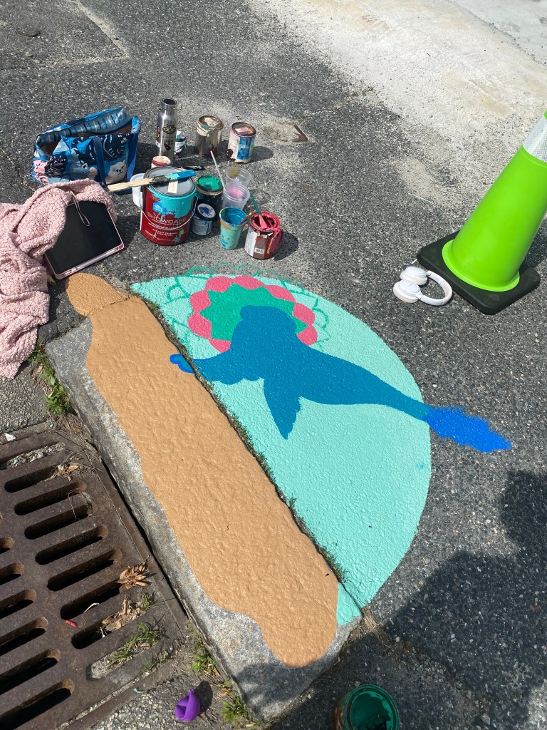 Artist painting the outline and background of the seal mural on the pavement. Blue, green, and pink colors are visible as the design takes shape.