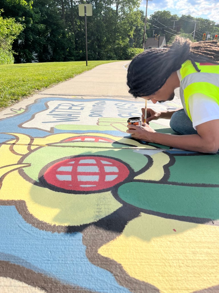 Artist Ryan Smith kneels down on the sidewalk in a yellow safety vest, using a small brush to paint details on the moth’s green body. The words of the mural are partially visible beneath him.
