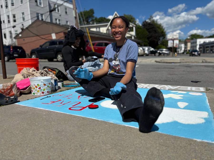 Savonne Pickett smiling sitting on the pavement while painting the “PURE WATER PURE JOY” mural. The mural is in progress, with red lettering and white cloud and flower shapes visible. Paint supplies, brushes, and a red bucket are scattered nearby under a sunny blue sky.