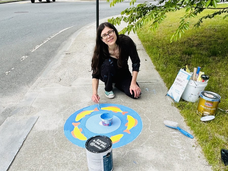 Artist Jill Strait kneeling on the sidewalk while painting a circular mural of yellow fish on a blue background. Paint cans, brushes, and tools are visible nearby.